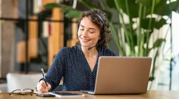 Eine Frau in gepunkteter Bluse sitzt mit einem Headset am Laptop und notiert etwas auf Papier